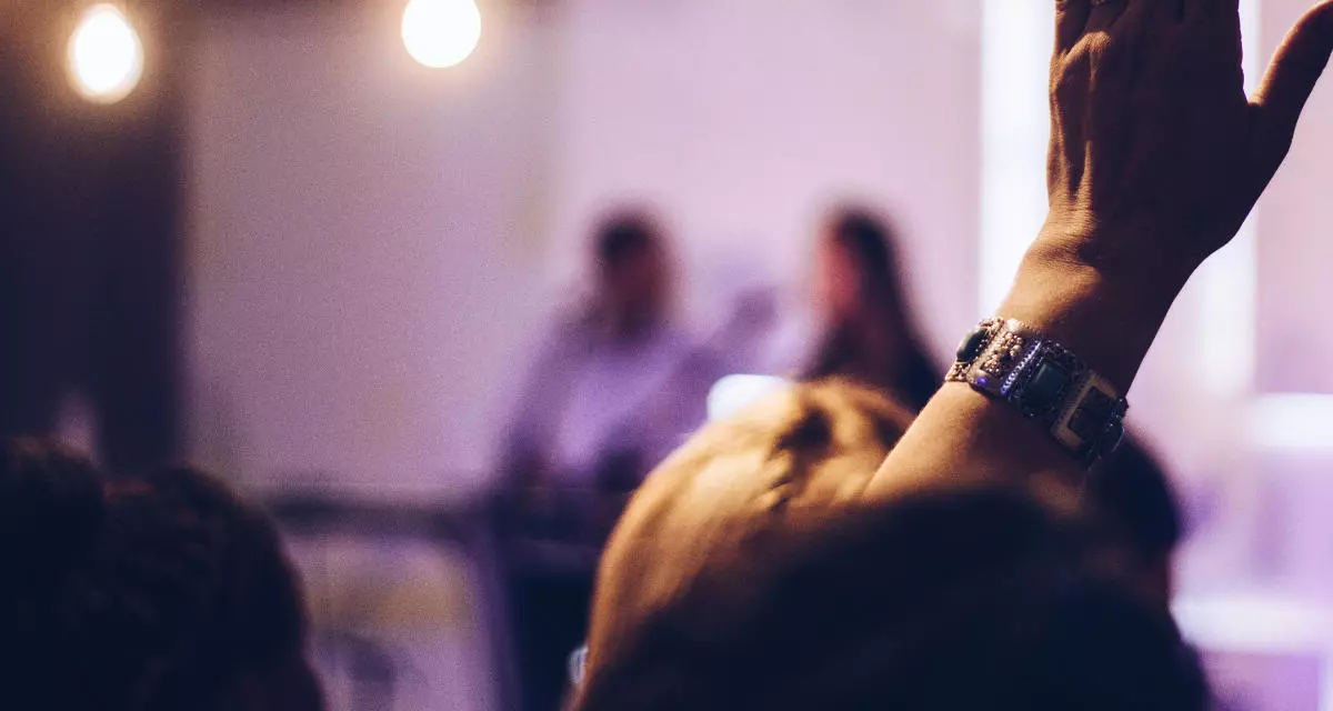 Symbolbild Veranstaltung: Marcos Luiz|Unsplash Menschen sitzen auf dem Podium, im Vordergrund eine erhobene Hand.