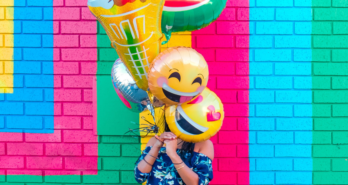 Mädchen vor farbiger Hauswand mit bunten Luftballons vor dem Gesicht
